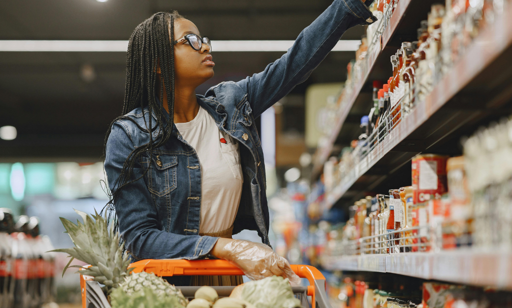 Mulher negra com tranças nos cabelo e óculos, usa uma jaqueta jeans e uma camiseta branca, ela está com uma luva transparente nas mãos, que se apoia ao carrinho de supermercado. Ela olha par a prateleira e o outro braço está levantado para pegar um produto ao alto.